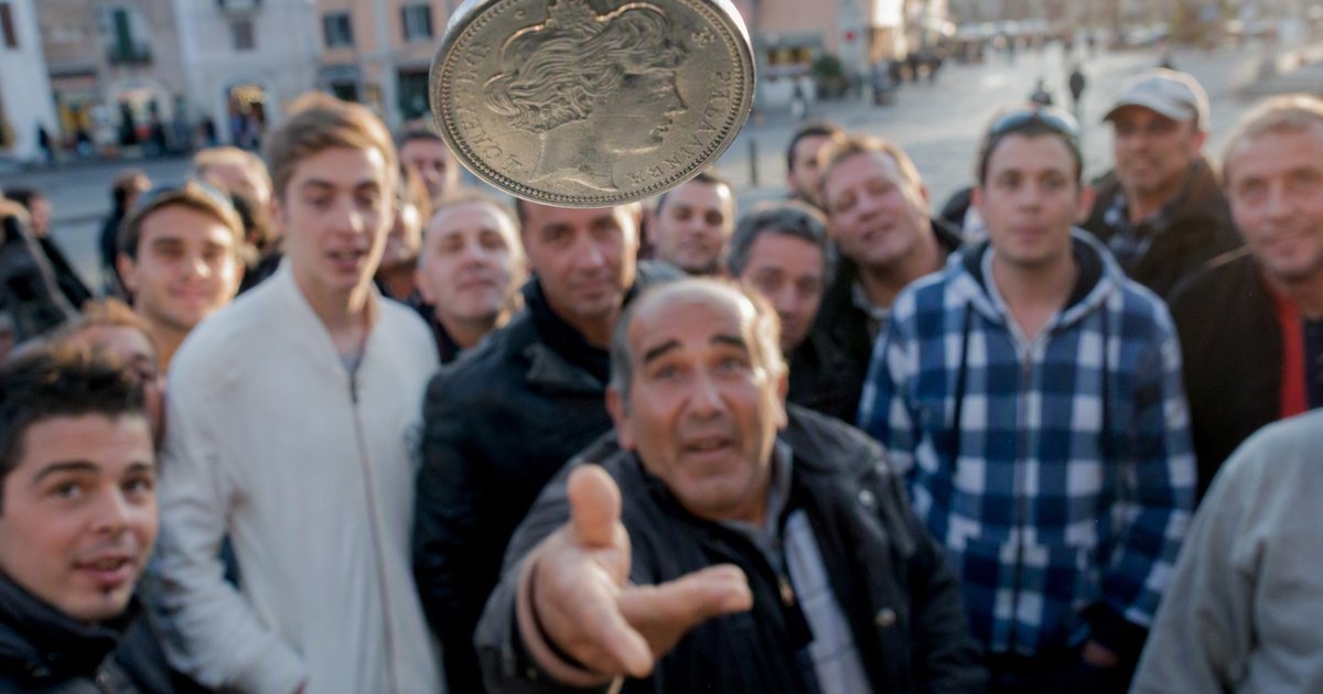 A large silver coin caught in mid-air in the foreground of a town square. Behind it a crowd looks up at the toss
