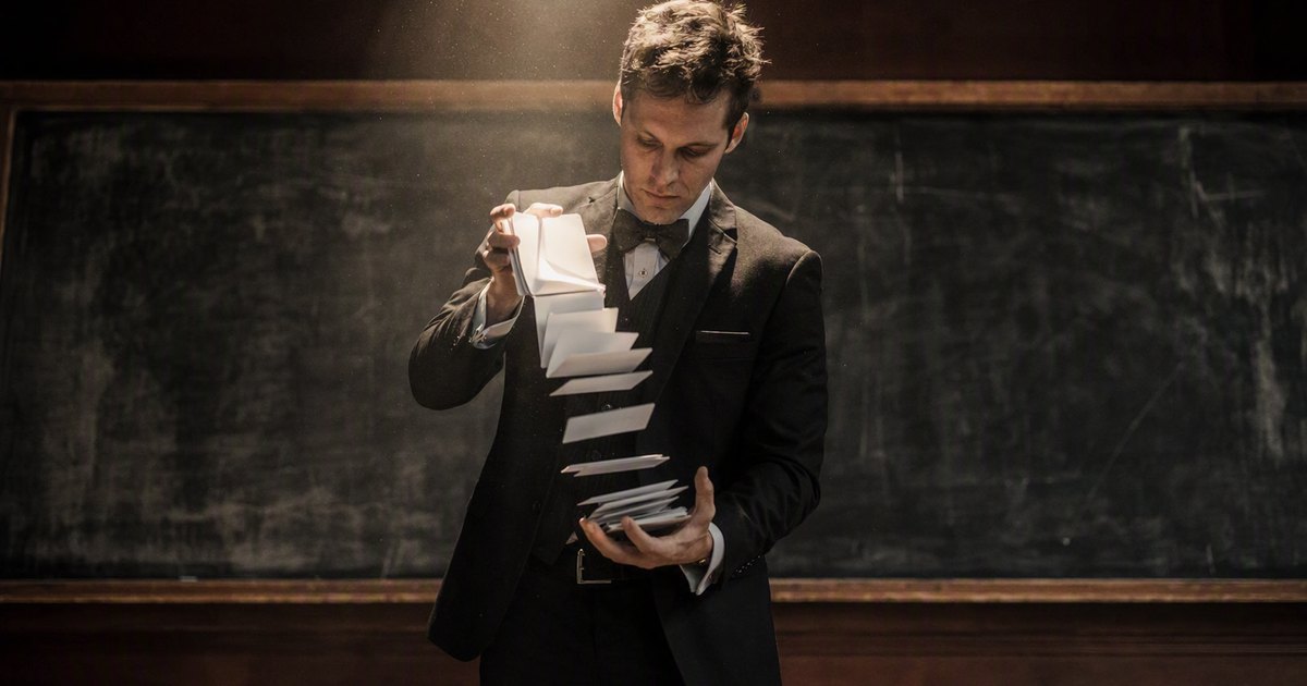 A stage magician shuffles an oversized deck of blank index cards under a spotlight. A chalkboard stands behind him